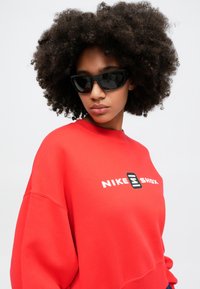 Červená mikina s čiernym logom "NIKE SHOX", čierne slnečné okuliare a kučeravé vlasy. Mäkká textúra a voľný strih.