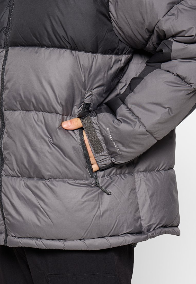 Chaqueta acolchada gris con una sección superior negra. Cuenta con bolsillos con cremallera, puños elásticos y una textura suave. Detalles del logotipo visibles.