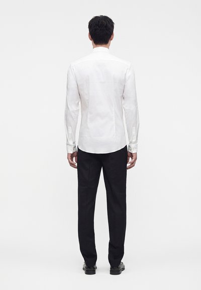 Man standing facing away wearing a fitted white long-sleeve shirt, black trousers, and black shoes against a plain white background.