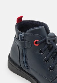 Marinblå läderankelstövlar med svart sula, med röda accenter, en sidodragkedja och rund snörning. Mjuk foder synlig inuti.