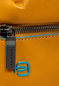 Geel leermateriaal met een matte afwerking, voorzien van een zwarte ritstrekker, blauwe accentstiksels en een blauw merkembleem.