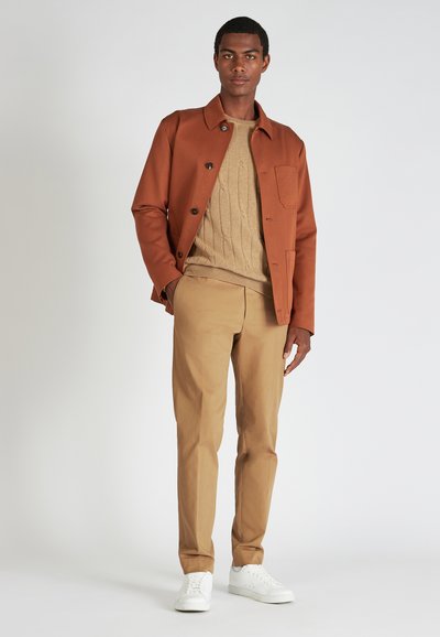 Chaqueta marrón con botones negros sobre un suéter de punto de cable color beige, combinada con pantalones marrones claros y zapatillas blancas. Diseño simple, textura suave.