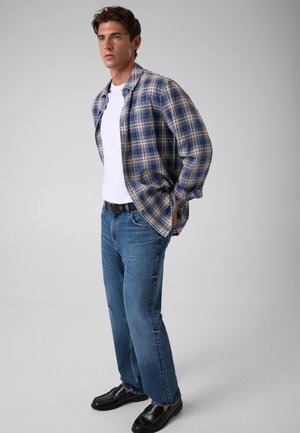 Ung mand iført blå og beige ternet skjorte over hvid t-shirt, blå jeans og sorte sko står mod en ensfarvet grå baggrund.