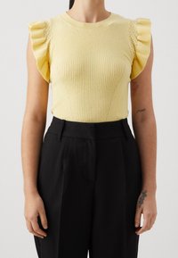 Kobieta w żółtej bezrękawnikowej bluzce z prążkowanej tkaniny z falbanami na ramionach oraz czarnych, plisowanych spodniach o wysokim stanie, na jednolitym tle.