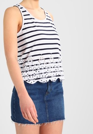Vrouw draagt een witte mouwloze tanktop met marineblauwe strepen en een bloemenkant zoom, gecombineerd met een blauwe denim minirok met rafelige randen.