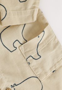 Beežist sametriidest kangas, millel on sinised karu joonised, volditud mansett ja tasku detail, rõhutades tekstuuri ja mängulist mustrit.