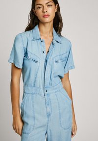 Light blue denim jumpsuit with short sleeves, front zip pockets, and a fitted waist. Features a button-up front and wide leg design.
