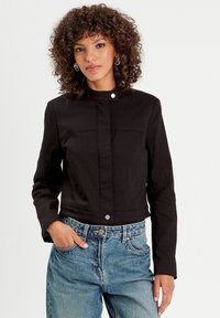 Black cropped jacket with a standing collar, front button closure, and two chest pockets. Worn over high-waisted denim jeans.