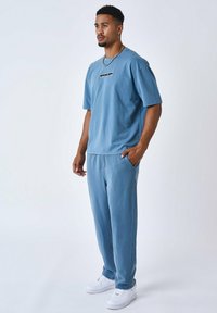Camiseta de algodón azul claro con un logo negro en el pecho, combinada con pantalones de chándal azules a juego y zapatillas blancas. Ajuste y diseño casual.