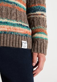 Suéter marrón de punto con rayas multicolores (naranja, azul verdoso, crema) que presenta un dobladillo acanalado y una etiqueta de tela en la parte inferior.