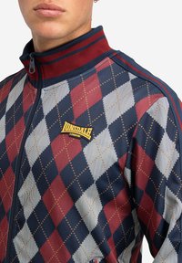 Navy, rode en witte argyle-patroon zip-up sportjack. Kenmerken zijn een hoge kraag, contrasterende strepen en een Lonsdale-logo op de borst.