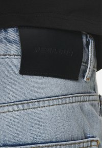 Jeans in denim blu chiaro con una toppa in pelle nera con la scritta "PEGADOR" in vita, indossati con una maglietta nera.