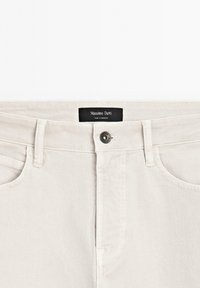 Světle béžové džíny z denimu s klasickým pětipocket designem, vybavené kovovým knoflíkem a zapínáním na zip, a s černou značkovou etiketou uvnitř.