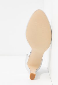 Chaussure à talon haut blanche avec une tige en cuir lisse et une semelle beige texturée. Le talon mesure environ 7,5 centimètres de haut et comporte un petit accent en caoutchouc.
