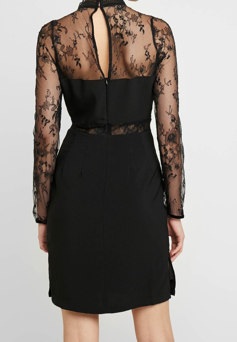 Femme portant une robe noire avec des manches longues en dentelle transparente, un panneau en dentelle dans le dos et une ouverture en forme de goutte d'eau dans le haut du dos.