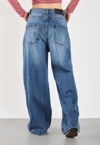 Weite blaue Jeans aus Denim mit einem verblassten Finish, mittelhoher Taille und zwei Gesäßtaschen. Der Stoff weist eine leichte Textur auf; der Saum ist genäht.