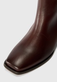 Brown leather ankle boot with a square toe, smooth texture, and minimalistic design. Features a black sole edge for contrast.
