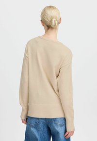 Maglione beige lavorato a maglia con maniche ampie, vestibilità rilassata e orlo a coste. La vista dal retro mostra un design semplice e una trama morbida. Sono visibili jeans in denim blu.