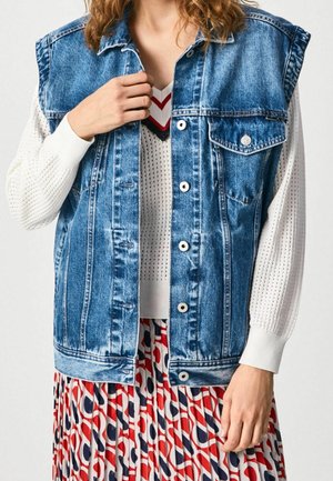 Mujer con chaleco vaquero azul sobre suéter blanco con estampado y falda plisada colorida en rojo, blanco y azul, sujetando el cuello del chaleco con una mano.