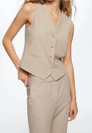 Gilet beige cintré sans manches boutonné, associé à un pantalon taille haute assorti, porté par une personne avec la main sur la hanche.