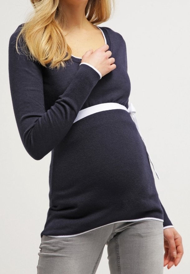 Femme enceinte portant un haut à manches longues bleu marine avec des bordures blanches et une ceinture blanche, associée à un pantalon gris clair.