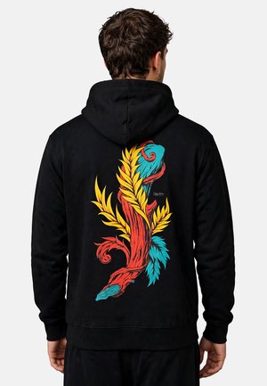 Hombre con sudadera negra con capucha con diseño abstracto de plantas y hojas en colores rojo, amarillo y azul en la espalda, de espaldas a la cámara.