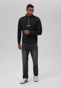 Svart hettejakke med høy krage, med en hvit logo på brystet. Brukt med mørke jeans og hvite sneakers.