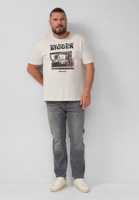 Beige graphic t-shirt met een vintage televisieprint; grijze jeans; witte sneakers met een lichtbruine zool; casual pasvorm, korte mouwen.