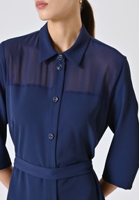 Marineblauwe jurk met een doorzichtige schouderpartij, knopenfront en een ceintuur op de taille. Bevat ronde knopen en driekwartmouwen. Soepele textuur.