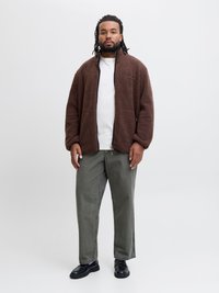 Veste en polaire marron et duveteuse avec fermeture éclair et poches latérales, portée sur un t-shirt blanc. Un pantalon gris et des chaussures noires complètent la tenue.