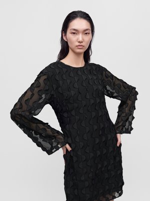 Un mannequin pose les mains sur les hanches, portant une robe noire texturée à manches longues transparentes, sur un fond gris uni.