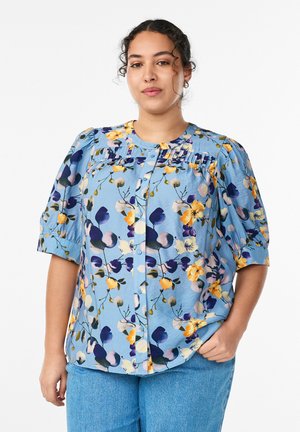 Kvinde med krøllet hår iført en blå blomsterknapbluse med puffede ærmer og lyseblå jeans, stående med den ene hånd i lommen.