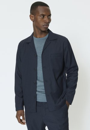 Marineblå skjortejakke med nålestriber, der har en reverskrave, knapper foran og en brystlomme, båret over en tekstureret lyseblå t-shirt.