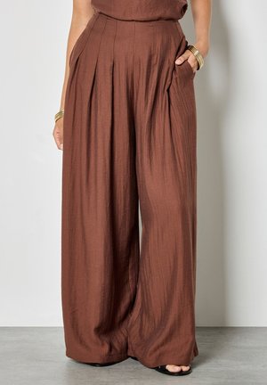 Frau trägt hoch taillierte, braune Hosen mit weitem Bein, Falten und seitlichen Taschen, kombiniert mit goldenen Armreifen und schwarzen Sandalen mit offenem Zehenbereich.