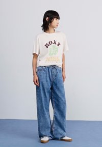 Loose-fitting blue denim jeans paired with a cream graphic T-shirt featuring Roxy branding and a green floral design.