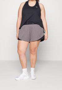 Persona vestida con una camiseta sin mangas negra, pantalones cortos deportivos grises, calcetines blancos y zapatillas blancas, de pie contra un fondo liso.