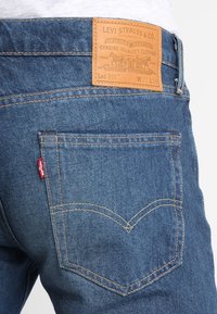 Modré džínové kalhoty s koženou nášivkou nesoucí nápis „Levi Strauss & Co.“ a zlatým prošitím. Mají zadní kapsu s charakteristickým šitím.