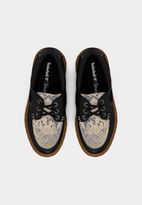 Paire de chaussures bateau Timberland noires et beige tachetées avec semelles marron et lacets noirs, vues de dessus sur un fond blanc.