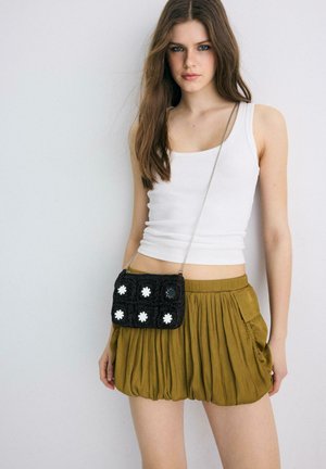 Giovane donna che indossa una canottiera bianca, una minigonna plissettata verde oliva e una piccola borsa nera a tracolla con motivi floreali bianchi.