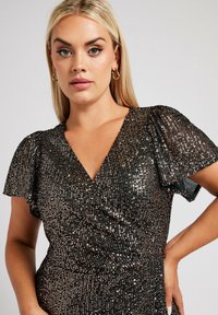 Czarna sukienka z cekinami, o dekolcie w kształcie litery V i falbankowymi rękawami; posiada dopasowaną talię. Błyszcząca, teksturowana tkanina tworzy olśniewający efekt.