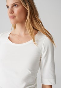 Blonde vrouw met blauwe ogen die een wit shirt met ronde hals en ellebooglange mouwen draagt, kijkt licht naar links.