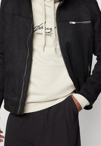 Chaqueta de ante negra con cierre de cremallera y bolsillos frontales, superpuesta a una sudadera beige con cordón y logo impreso.