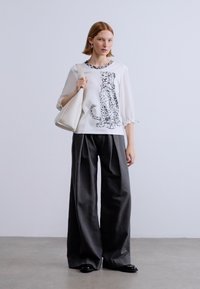 Λευκό μπλουζάκι με τύπωμα γάτας, κοντά φουσκωτά μανίκια· συνδυασμένο με σκούρο παντελόνι φαρδύ. Το μοντέλο κρατά μια μπεζ τσάντα και φοράει μαύρα παπούτσια.