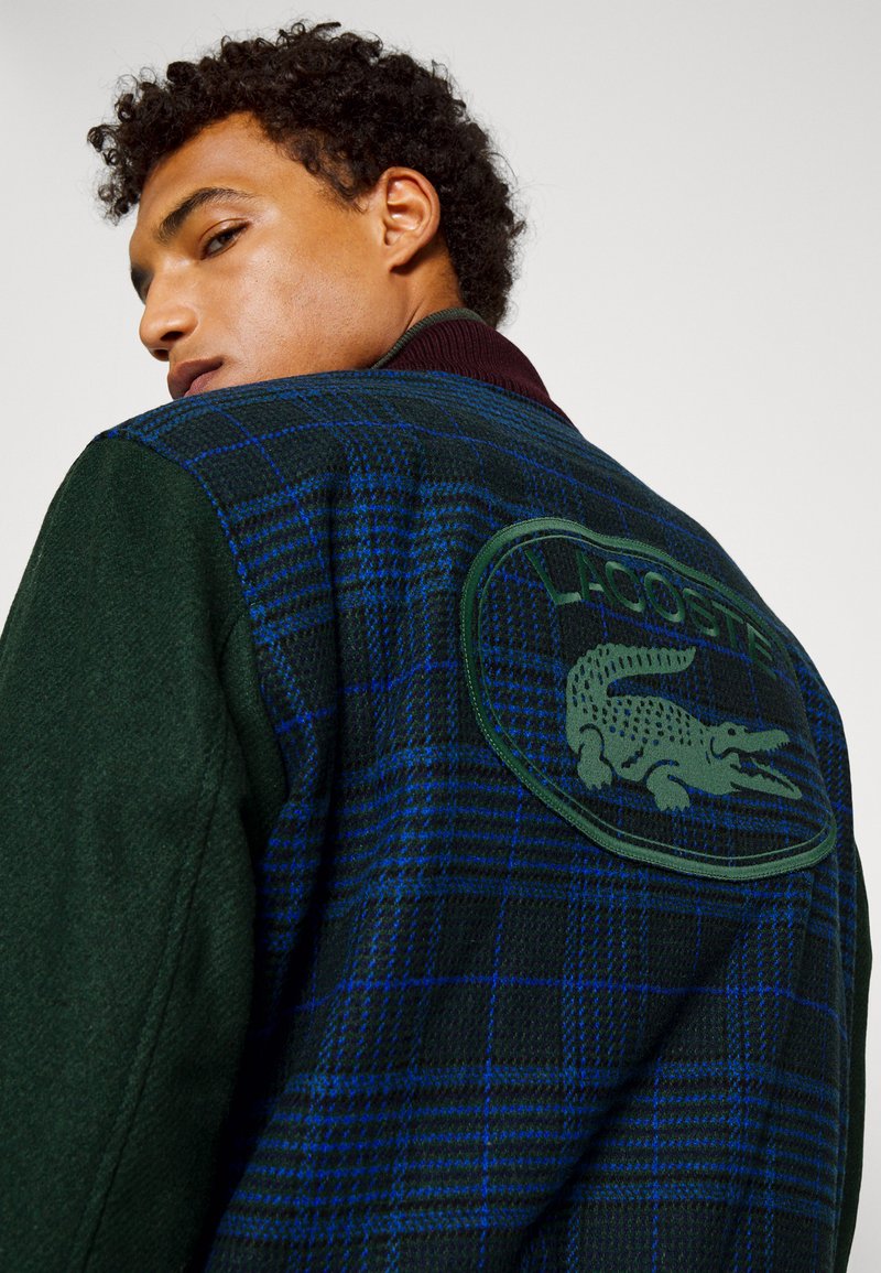 Veste en tartan vert et marine avec un col bordeaux, dotée d'un grand écusson logo Lacoste vert à l'arrière. Texture douce et mélange de laine.