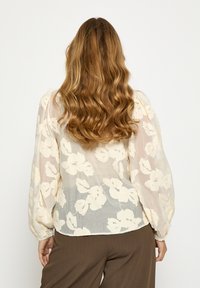 Lekka, kremowa bluzka z teksturowanym kwiatowym wzorem, bufkami i elastycznymi mankietami. Przepływający, półprzezroczysty materiał.