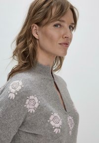 Grau gestrickter Pullover mit hohem Kragen, ausgestattet mit pinken Blumenstickereien auf der Vorderseite. Weiche Textur, leichtes Design und eine Schlüssel-Ausschnitt.