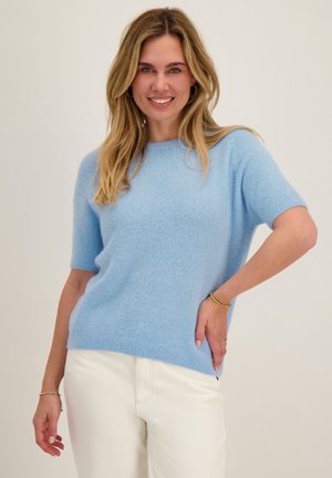 Glimlachende vrouw met blond haar, die een lichtblauwe korte mouwen trui en een witte broek draagt, staand met één hand op haar heup.