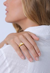 Guld ring med en rund klar ædelsten, der har et organisk design. Modellerens hånd hviler på en hvid, tekstureret stofbluse.