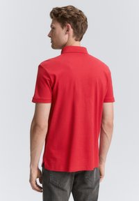 Rød polo t-shirt med korte ærmer og krave, der har en glat tekstur. Bæres med mørkegrå jeans, der viser en afslappet pasform.