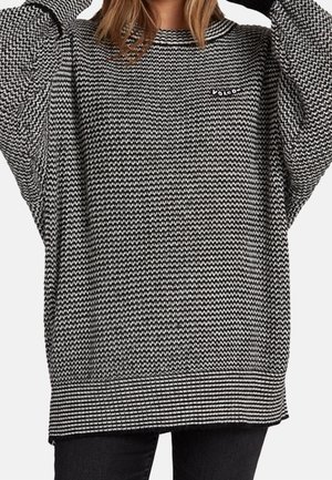 Femme portant un pull surdimensionné à motifs en zigzag noir et blanc avec un col rond et un petit logo sur la poitrine, associé à un pantalon noir.